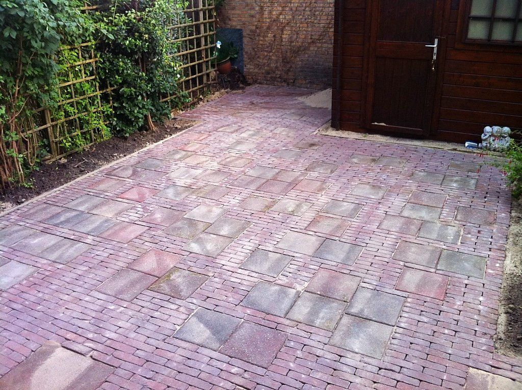 Een stenen terras in een tuin, dat overgaat in een cirkelvormig patroon, naast een houten schuur en groen.