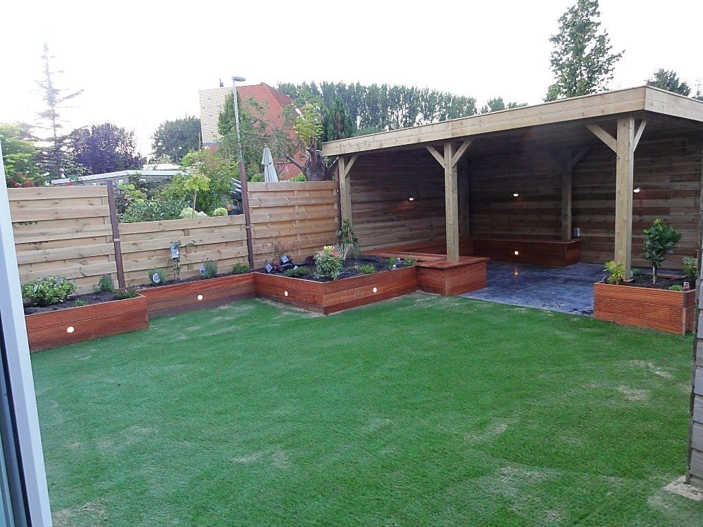 Een achtertuin met een houten schutting, verhoogde bloembedden en een overdekt terras. Groen gras en een blauwe lucht.