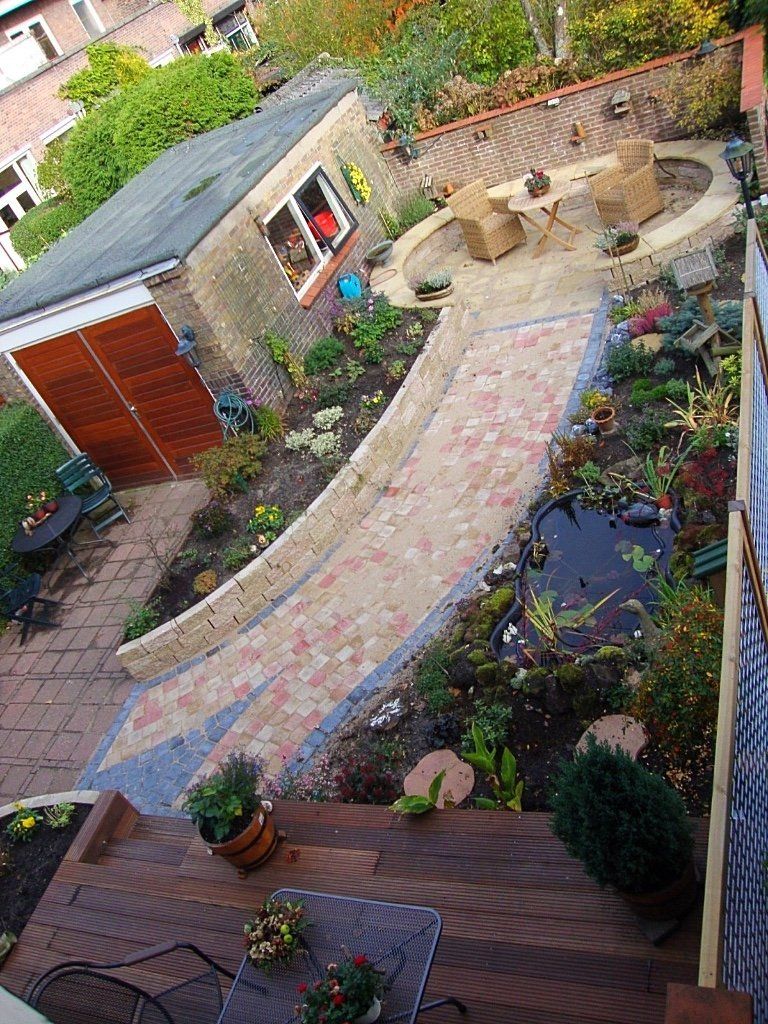 Een bakstenen pad slingert door een tuin en leidt naar een zitgedeelte. Links bevindt zich de garage, rechts de vijver.