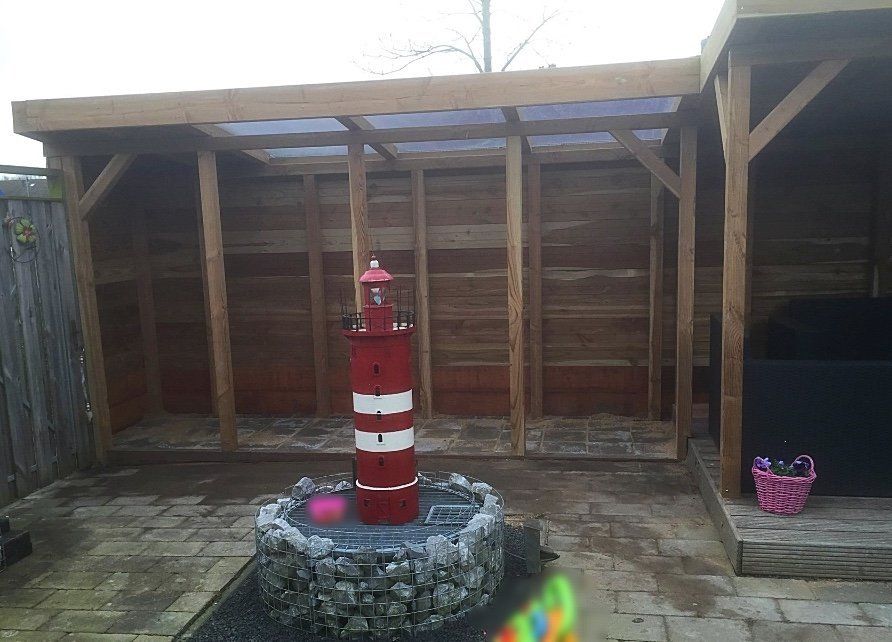 Houten schuur met een rood-wit vuurtorenbeeld op een stenen sokkel, op een bakstenen terras.