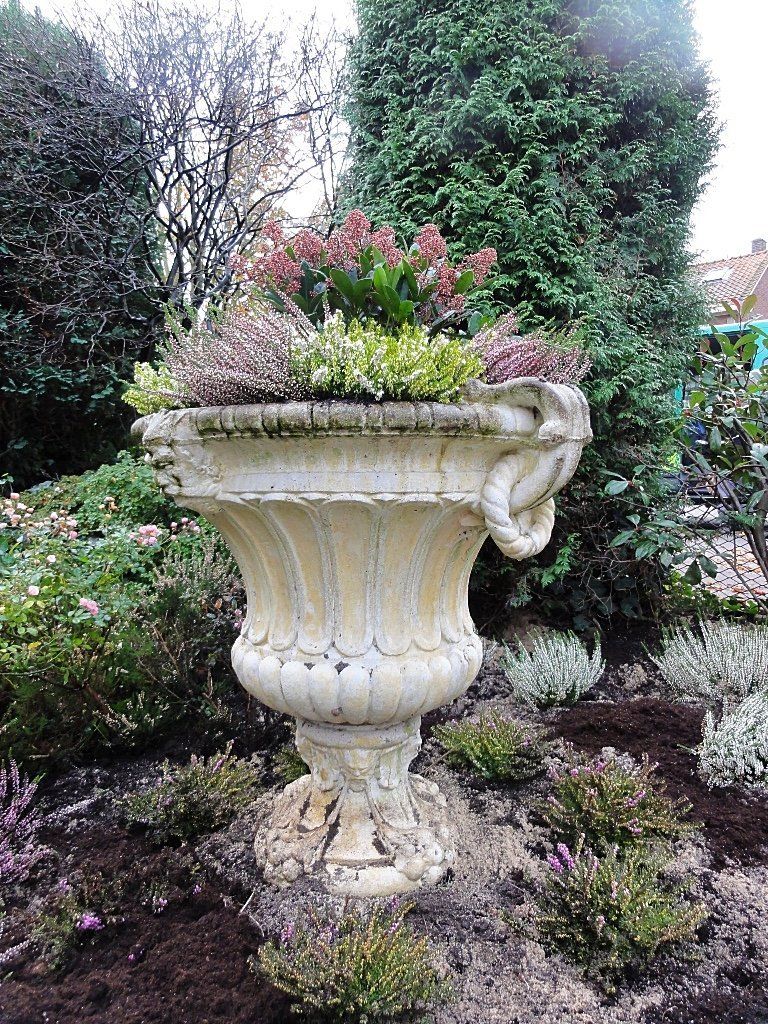 Een stenen plantenbak gevuld met planten in een tuin, met groenblijvende bomen op de achtergrond.