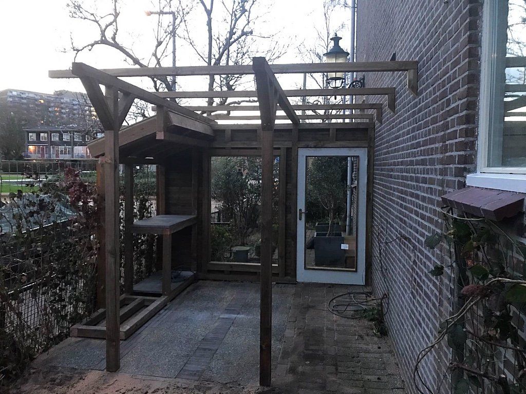 Houten pergola aan een bakstenen gebouw; deur en raam zichtbaar. Grijs terras. Bewolkte lucht.