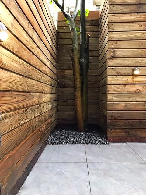 Afgesloten patio met houten lambrisering, grijze tegelvloer, boom en grind als bodembedekking.