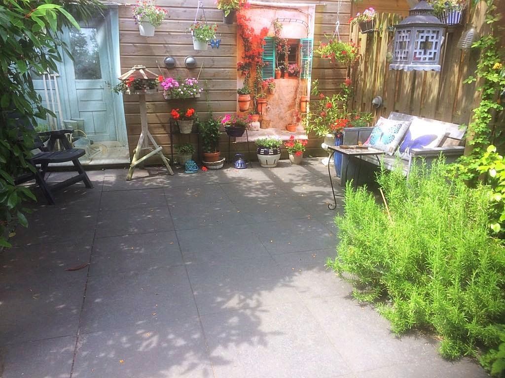 Terras in de achtertuin met bloemen, planten en een barbecue, versierd met hangende bloembakken en een lantaarn.