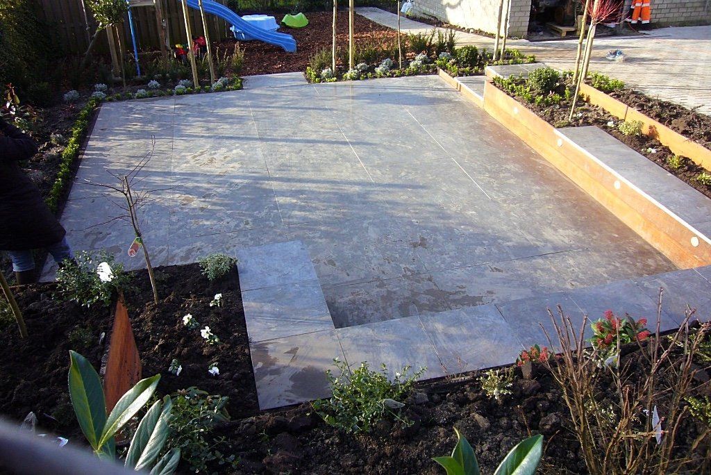 Een betonnen terras in een tuin, omgeven door bloemperken en houten borders.