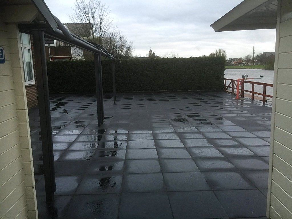 Een nat, donker betegeld terras met een zwarte overkapping. Een dichte groene haag vormt de achtergrond onder een bewolkte hemel.