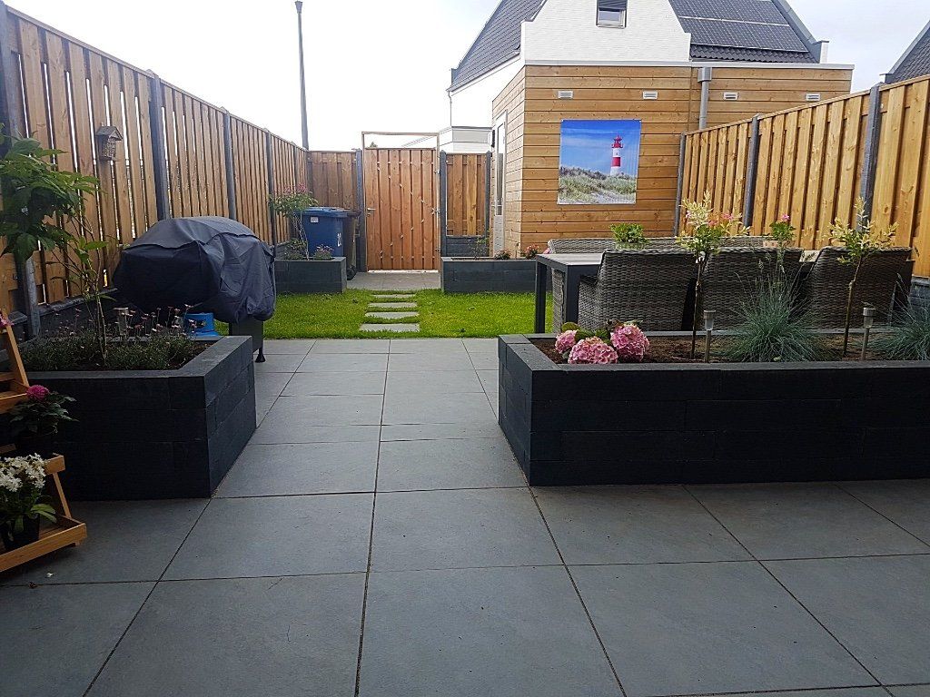 Een terras in de achtertuin met donkergrijze tegels, zwarte plantenbakken en een houten schutting.