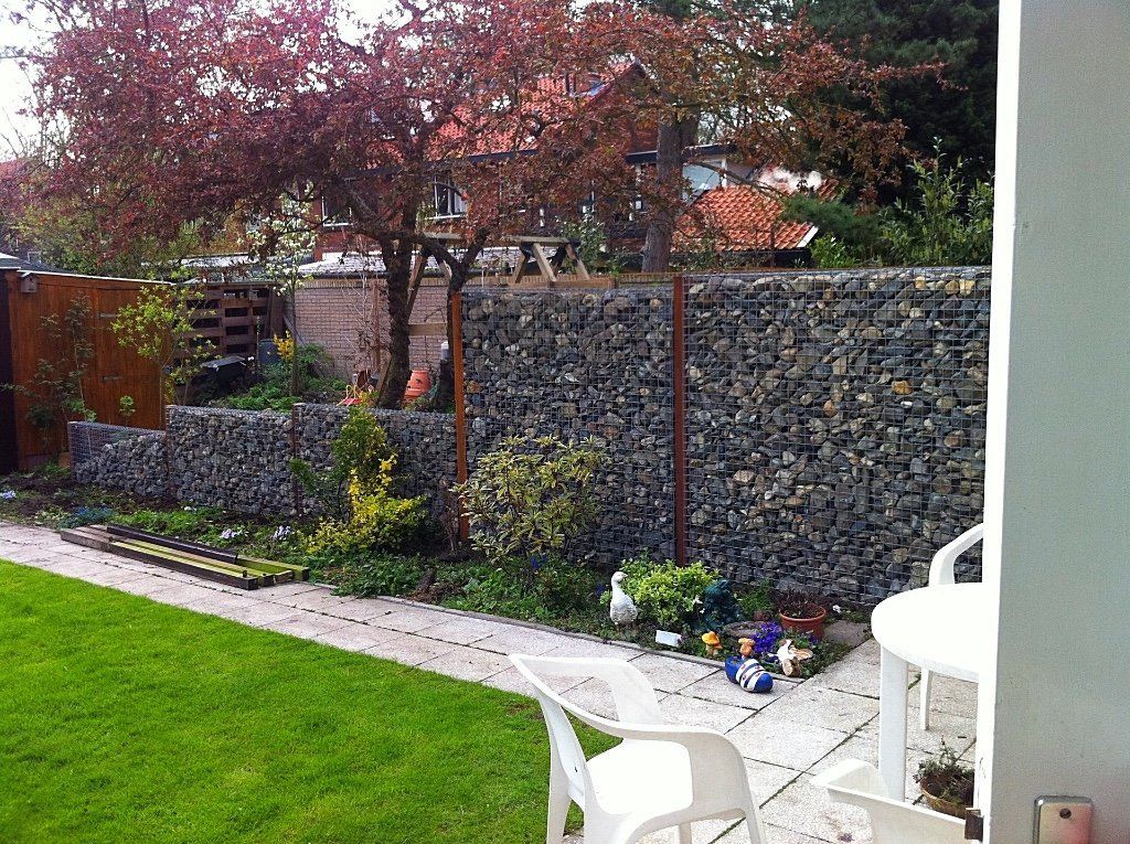 Een tuin met een met stenen gevulde gabionomheining, een gazon, bomen met rode bladeren en wit tuinmeubilair.