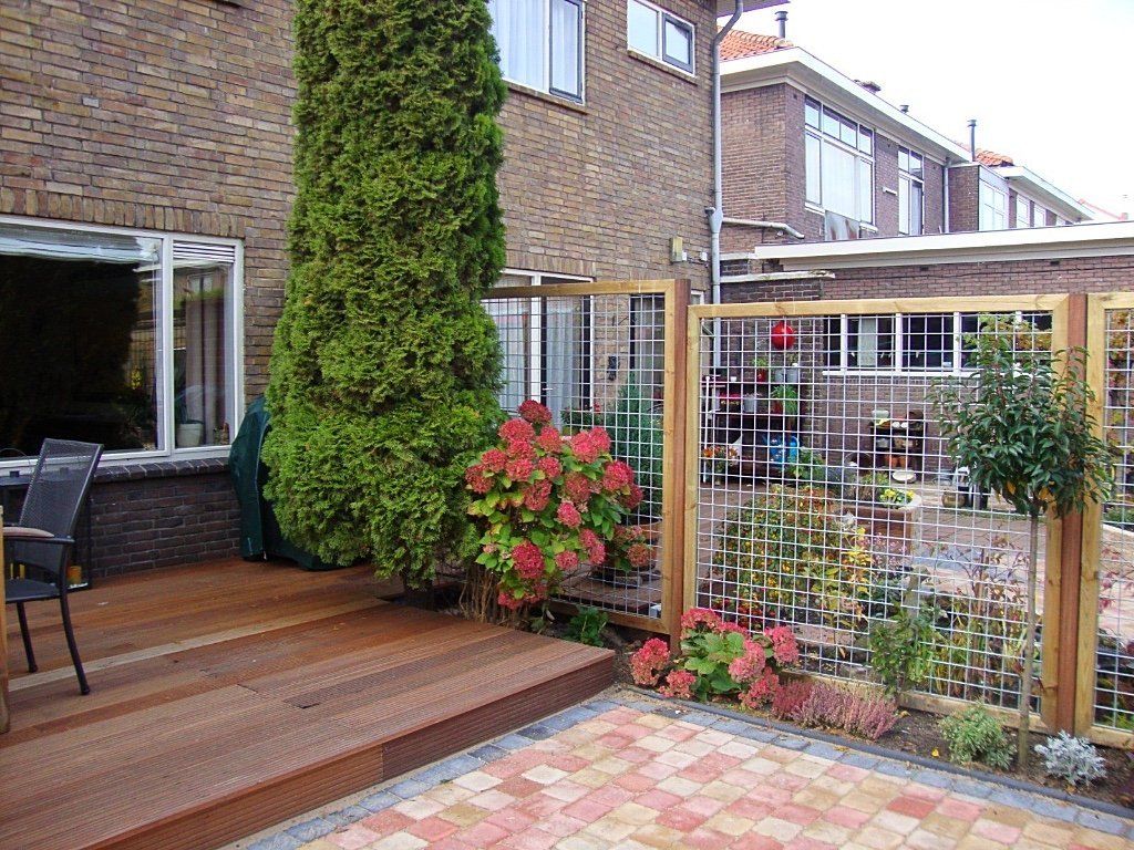 Achtertuin met een houten vlonder, een stenen patio, groene planten en decoratieve houten schermen.