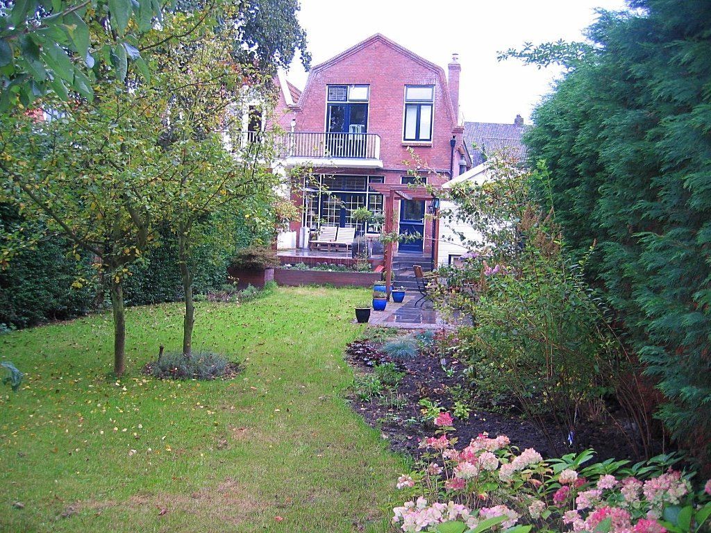 Achtertuin met groen gazon, bomen, bloemen en een bakstenen huis.