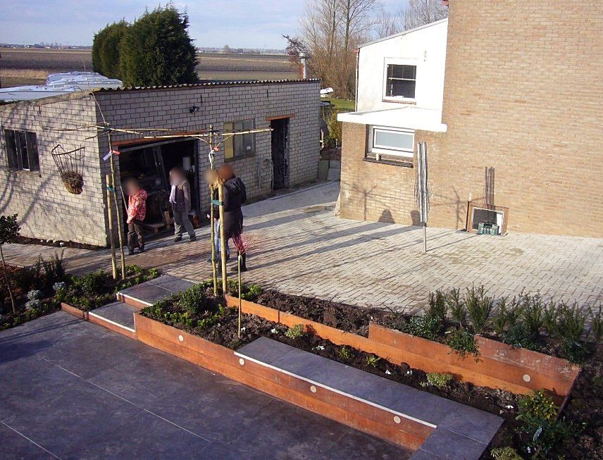 Achtertuin met plantenbakken in verschillende niveaus, mensen bij een garage en een bakstenen huis.