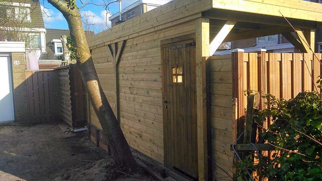 Houten schuur met deur en klein raam naast een boom en een hek.