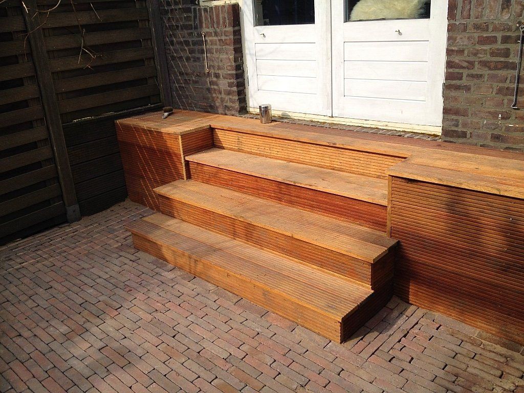 Houten treden leiden naar witte dubbele deuren, die zich bevinden op een stenen terras tegen een stenen muur.