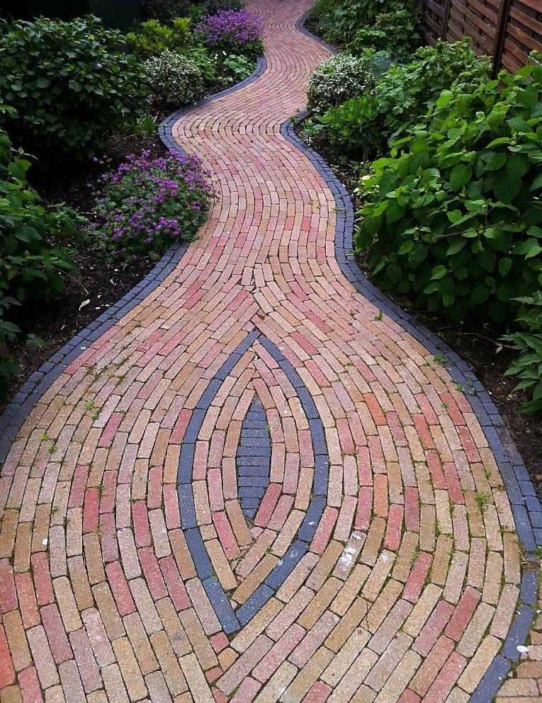Een bakstenen pad slingert door een tuin, met een oogvormig ontwerp in het midden. Weelderige groene struiken omzomen de zijkanten.