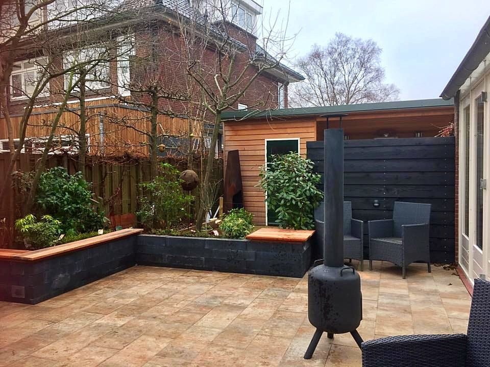 Terras in de achtertuin met bloembedden, zwarte schutting, een schuur en zitgelegenheid.