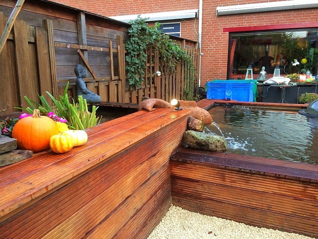 Een vijver met houten rand, pompoenen, een waterpartij en planten in een achtertuin.
