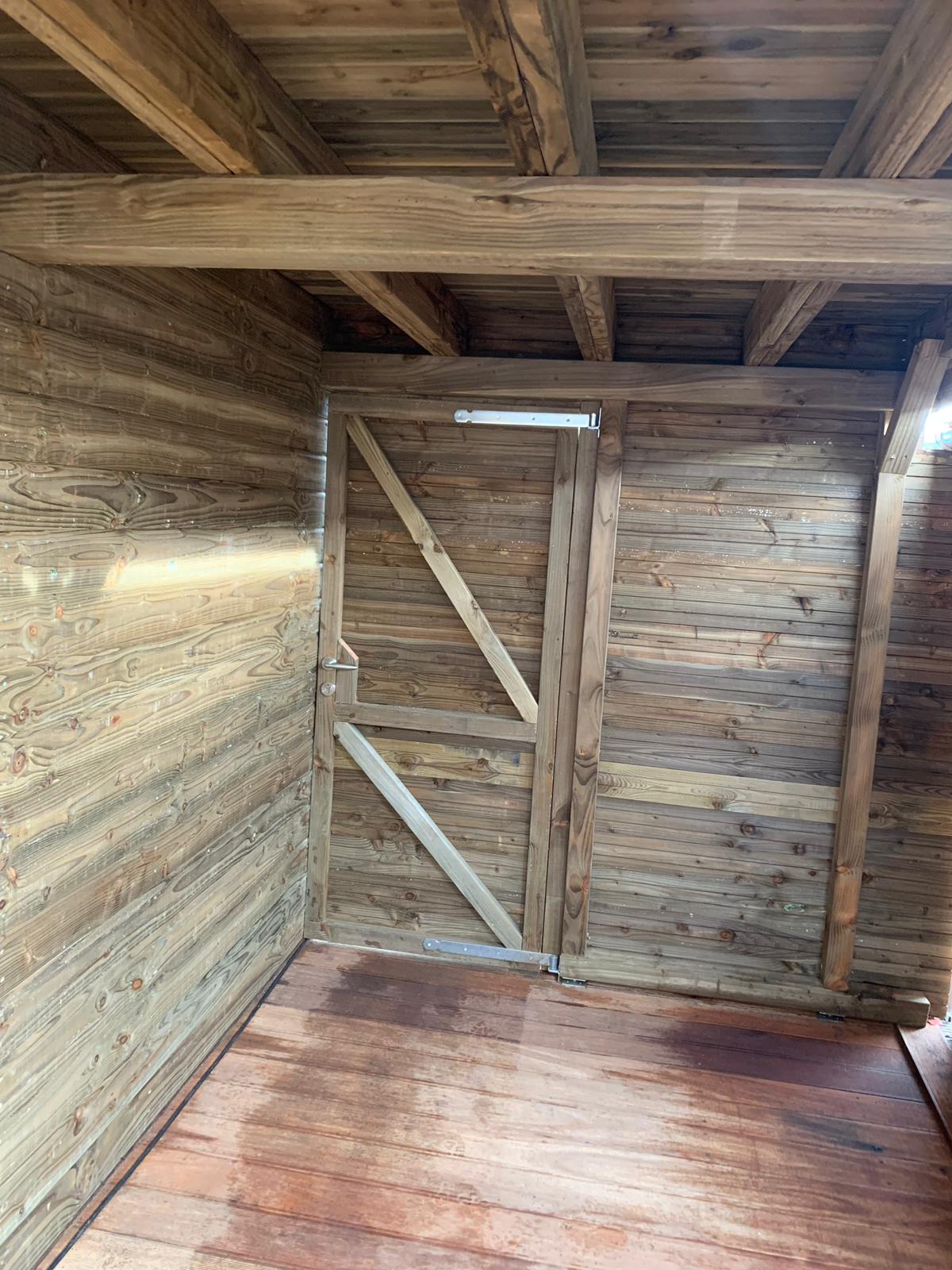 Interieur van een rustieke houten constructie. Zichtbaar zijn de muren, plafondbalken en een eenvoudige houten deur.