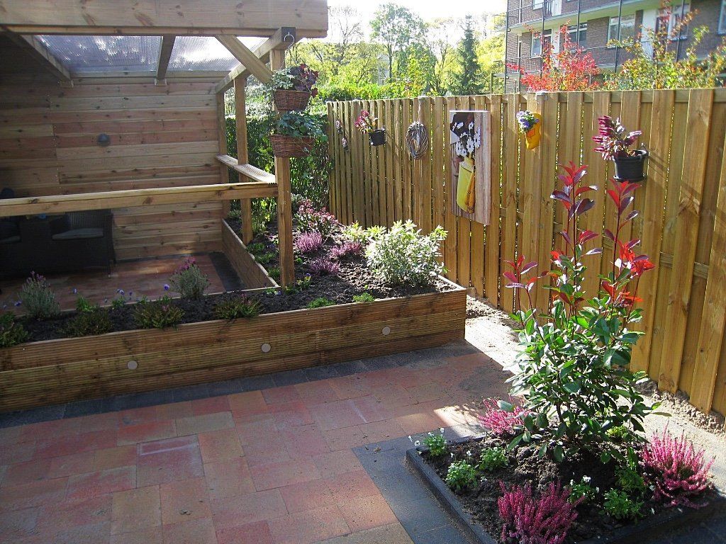 Een zonnig terras in de achtertuin met een houten schutting, verhoogde bloembedden en bloeiende planten.