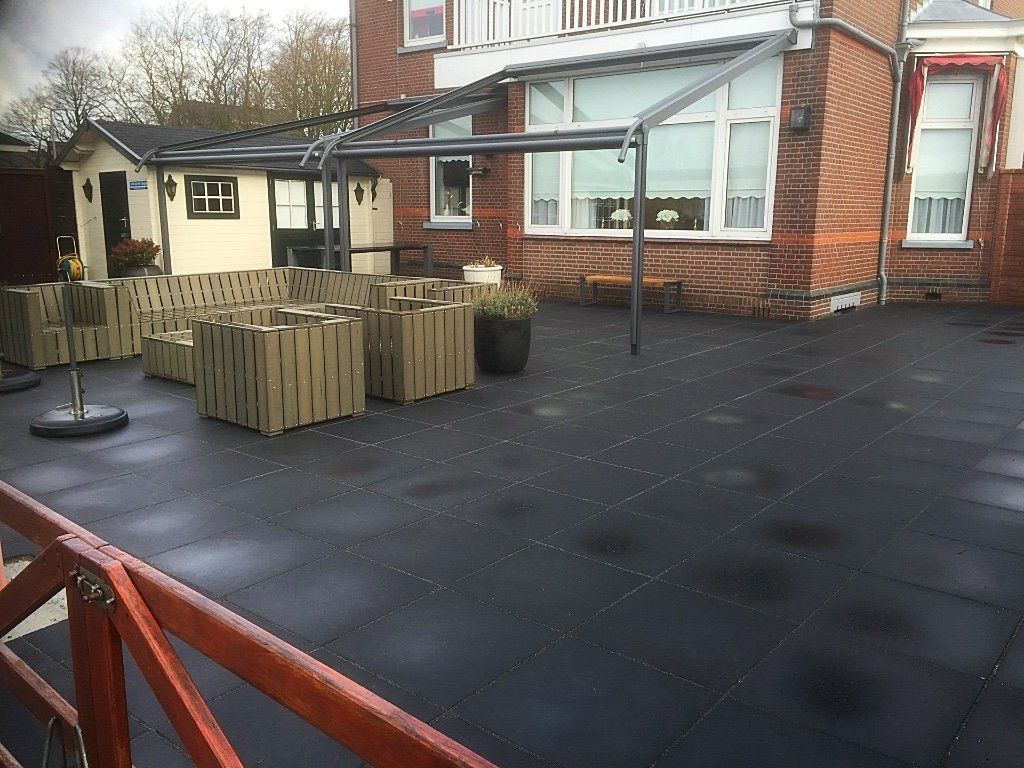 Terras met zwarte tegels, zitgelegenheid, overkapping en een bakstenen gebouw met ramen.