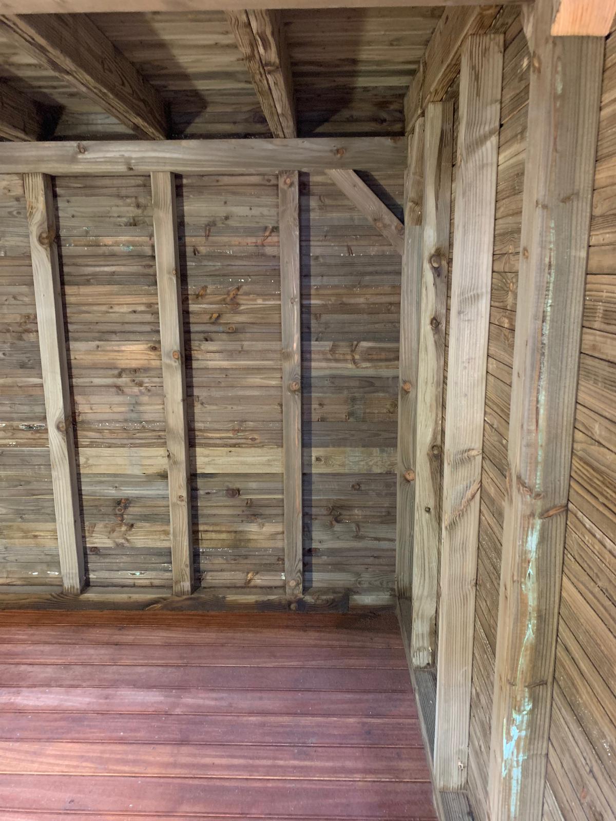 Interieur van een houten constructie: wanden van planken, balken en een roodachtige vloer.