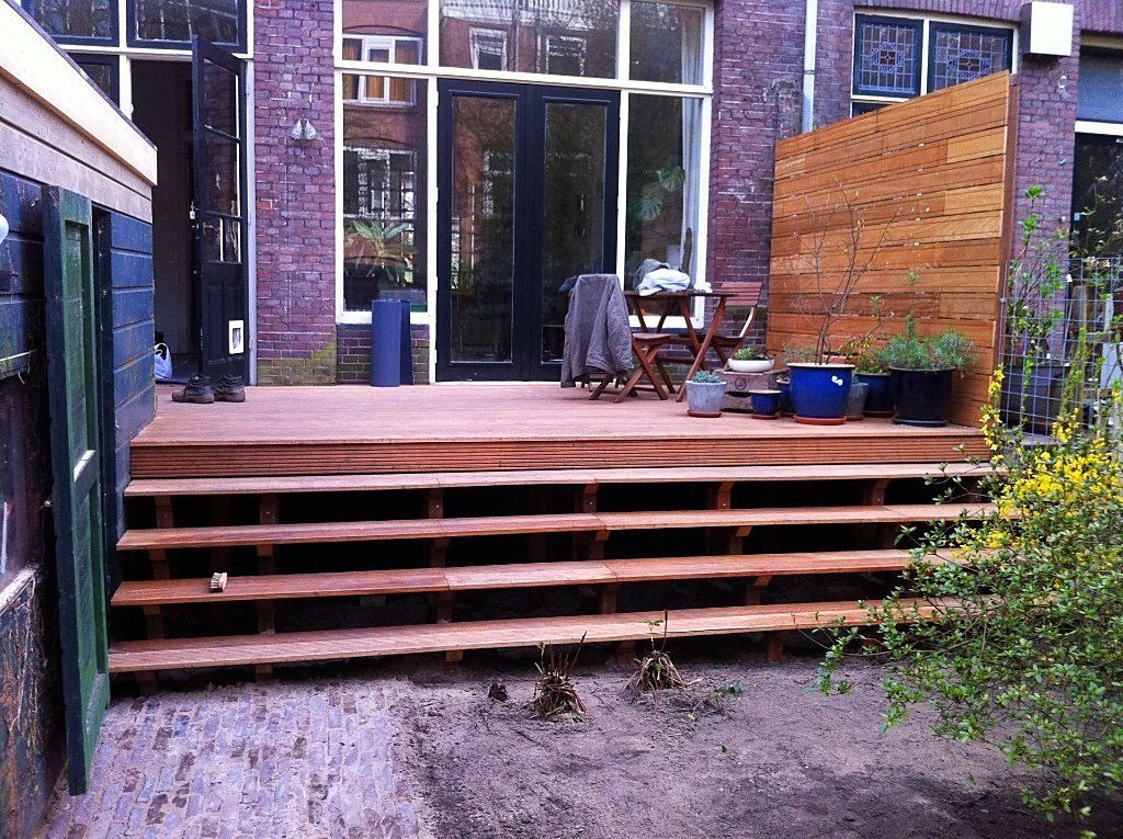Houten terras met trappen in een achtertuin, dat leidt naar een bakstenen gebouw met een deur en ramen.