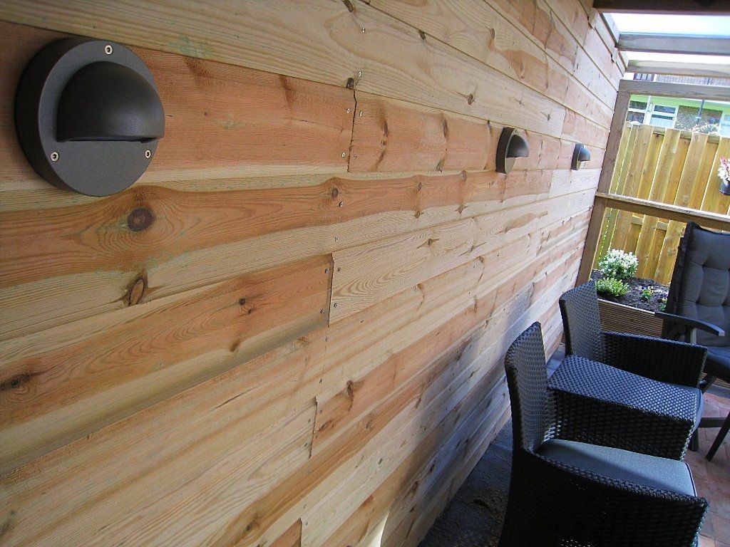 Houten buitenmuur met drie donkere wandlampen en terrasstoelen.