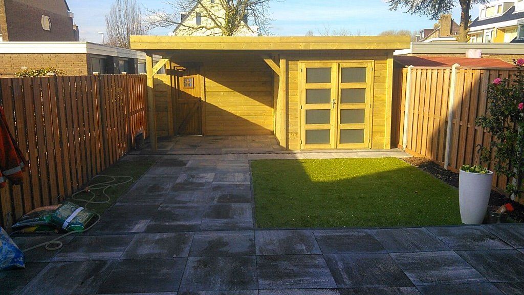 Een achtertuin met een houten schuur, terras en schuttingen; groen gras en zonlicht.