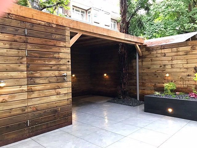 Houten schuur met openslaande schuifdeur op een betegeld terras; kleine lampjes, plantenbakken en een gebouw zijn zichtbaar.