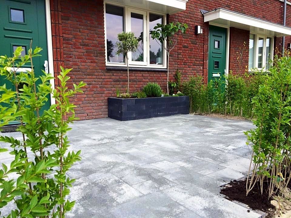 Entree van een huis van baksteen met grijze bestrating, groene deur en potplanten.