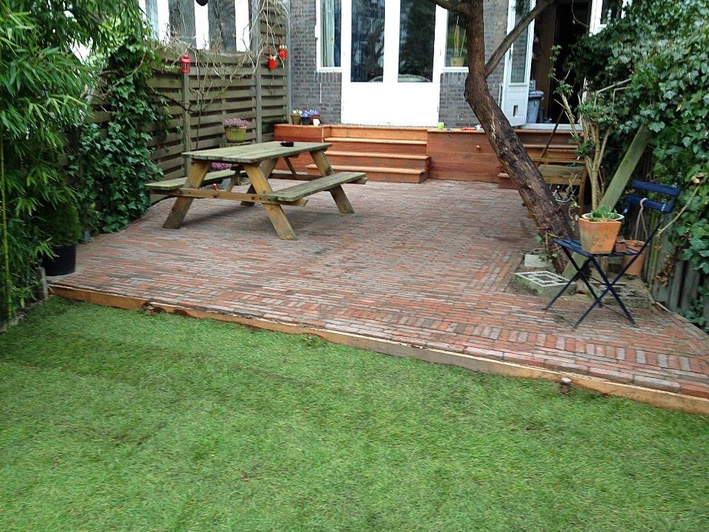 Een stenen terras met picknicktafel en een grasveld, houten trappen leiden naar een witte deur.
