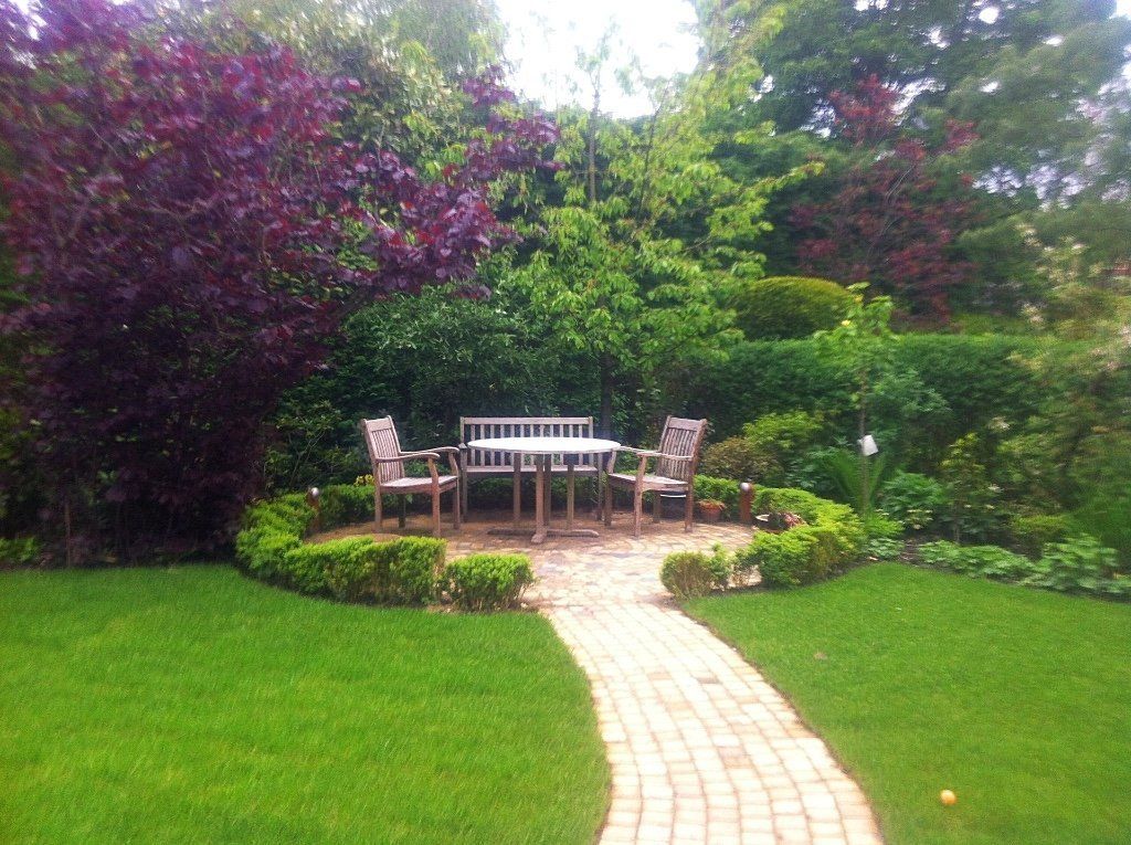 Een weelderige tuin met een bakstenen pad dat leidt naar een tafel en stoelen, omgeven door groen en een keurig gesnoeide heg.
