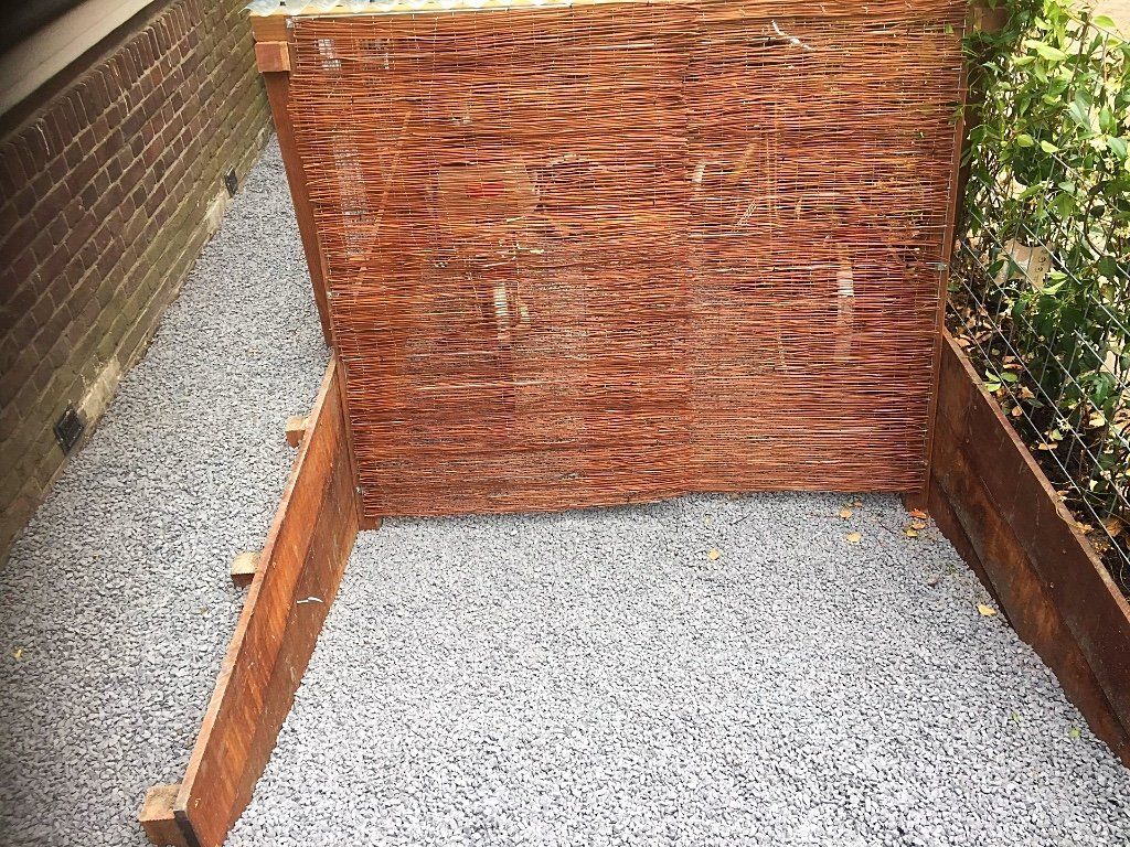 Houten compostbak met open deksel, geplaatst in grind tegen een bakstenen muur en begroeiing.