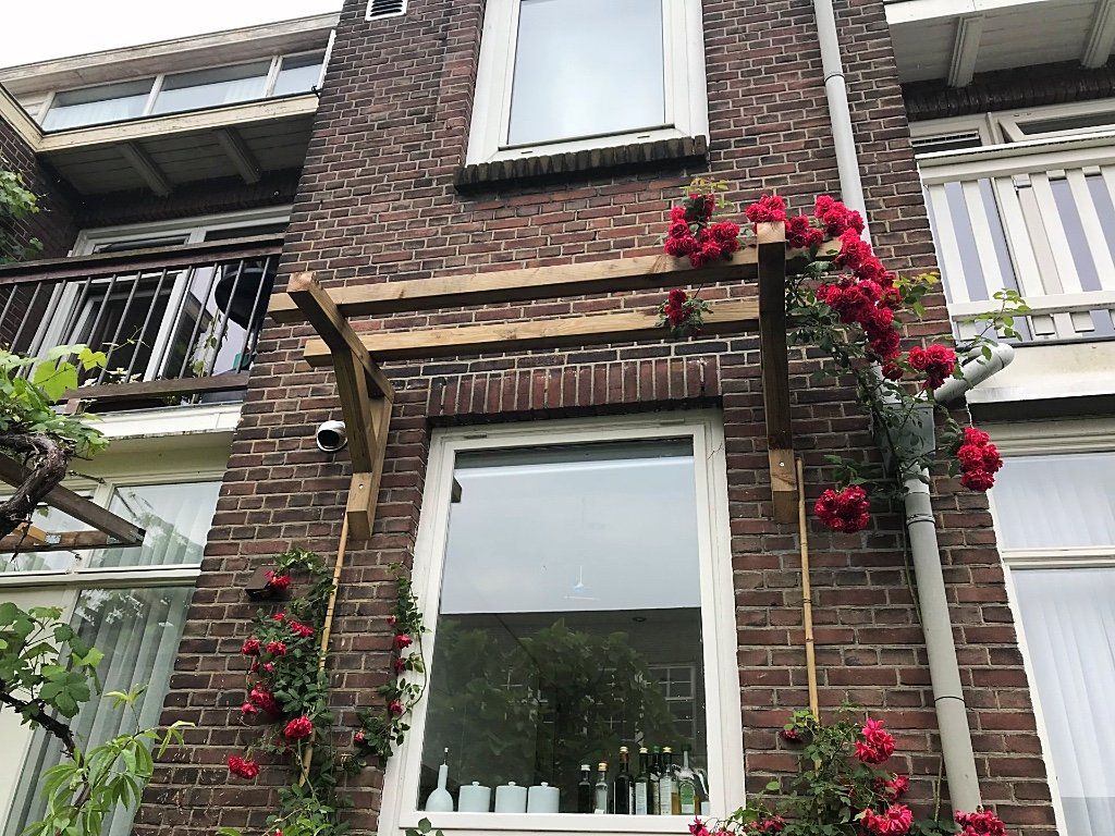 Bakstenen gevel met raam begroeid met klimrozen en houten pergola.