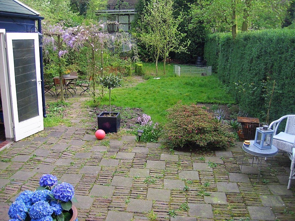 Achtertuin met een stenen terras, een klein gazon, bloeiende struiken en een schuur met een open deur.