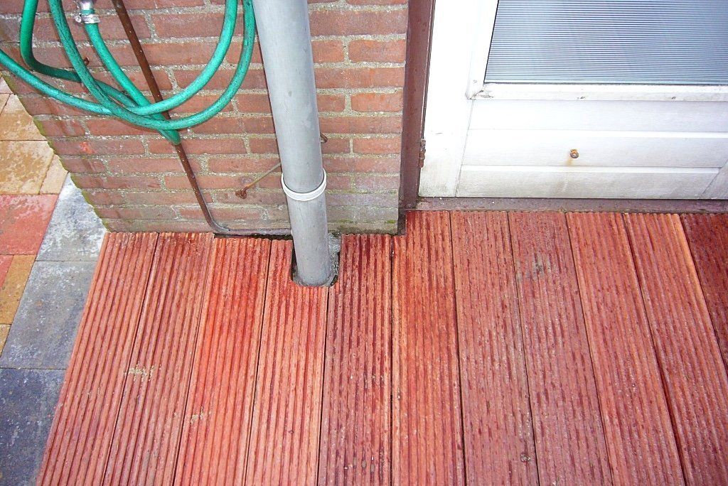 Een terras van rood hout met een grijze regenpijp, een bakstenen muur en een groene tuinslang.