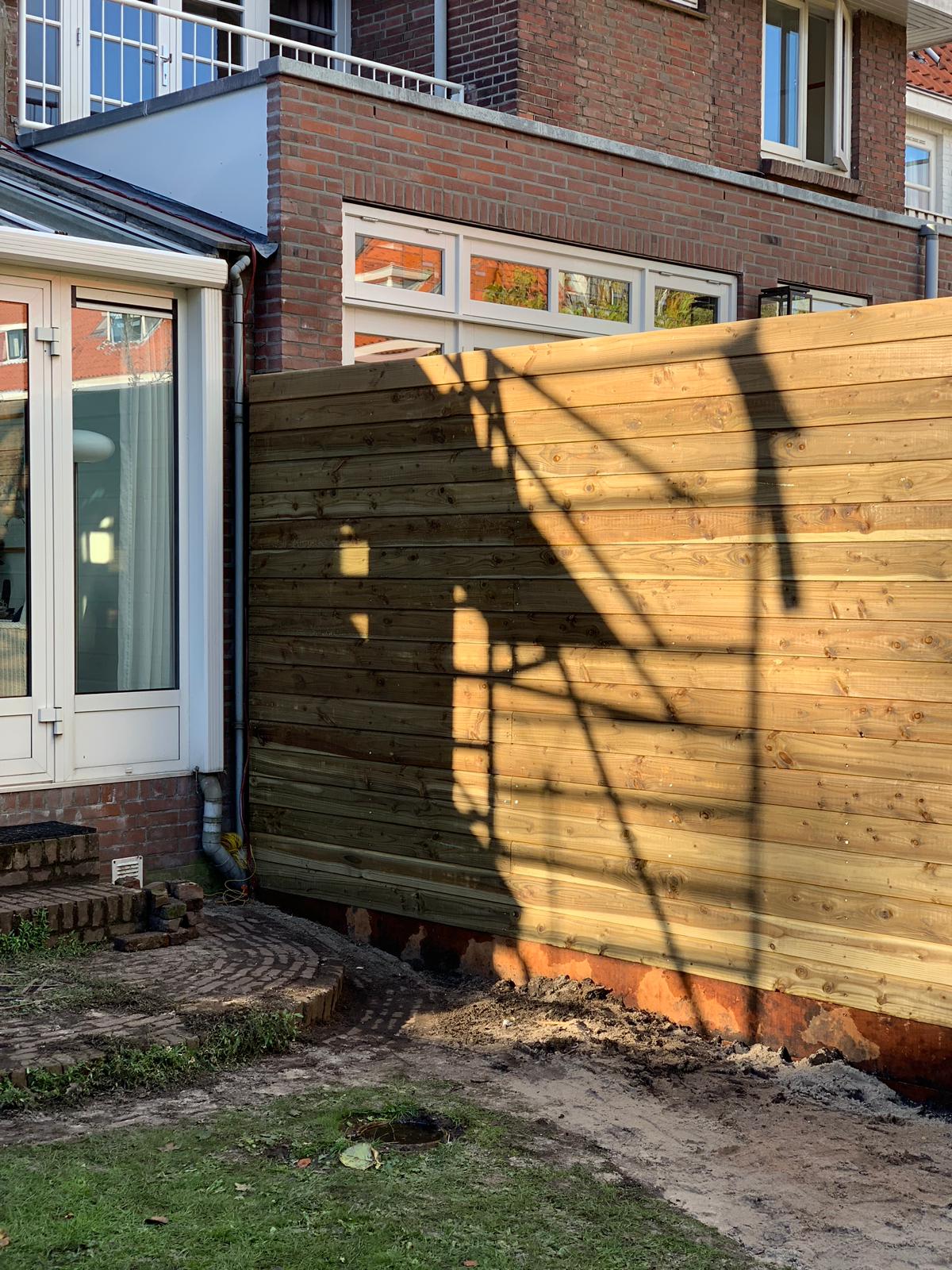 Een houten schutting naast een gebouw werpt een schaduw. Zonlicht verlicht de schutting.
