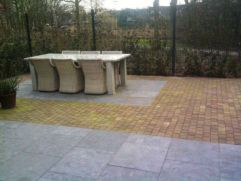 Terras met grijze natuurstenen tegels, een bakstenen rand en een rieten tafel met stoelen. Op de achtergrond een schutting en begroeiing.