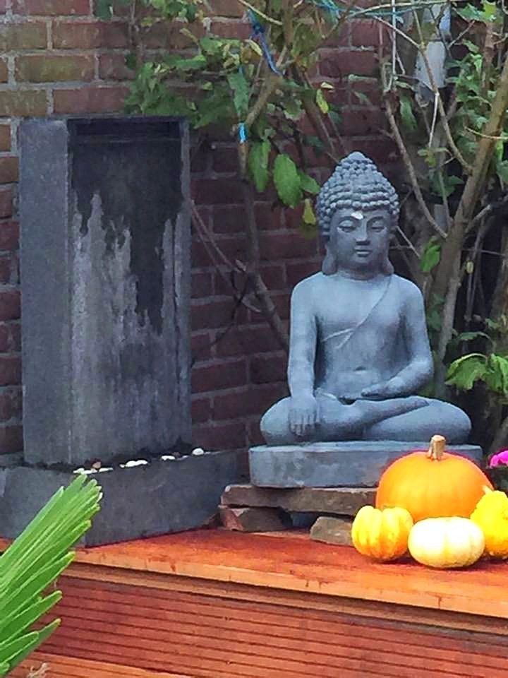 Boeddhabeeld bij een stenen fontein met pompoenen op een houten ondergrond, met een bakstenen muur op de achtergrond.