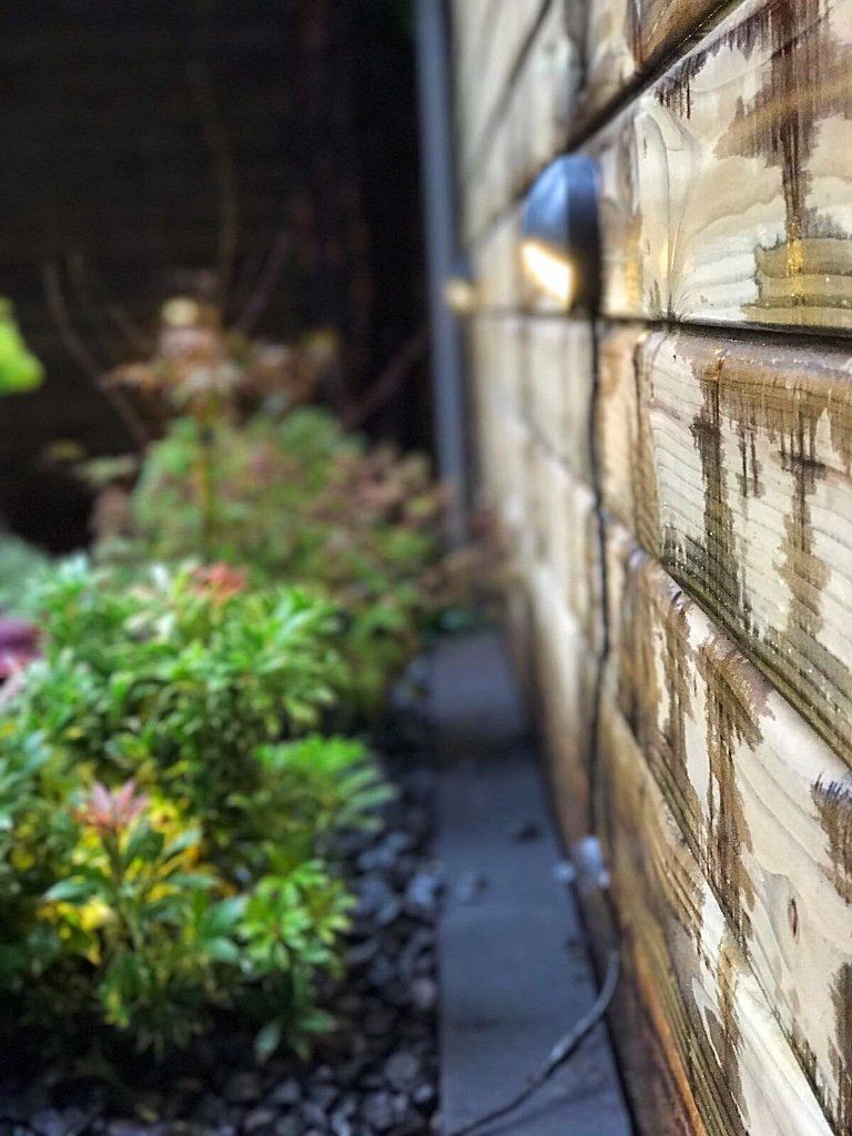 Een gebouw met houten lambrisering aan de buitenkant, met een spotlicht op een perk van groene en rode struiken en zwarte stenen in een tuin.