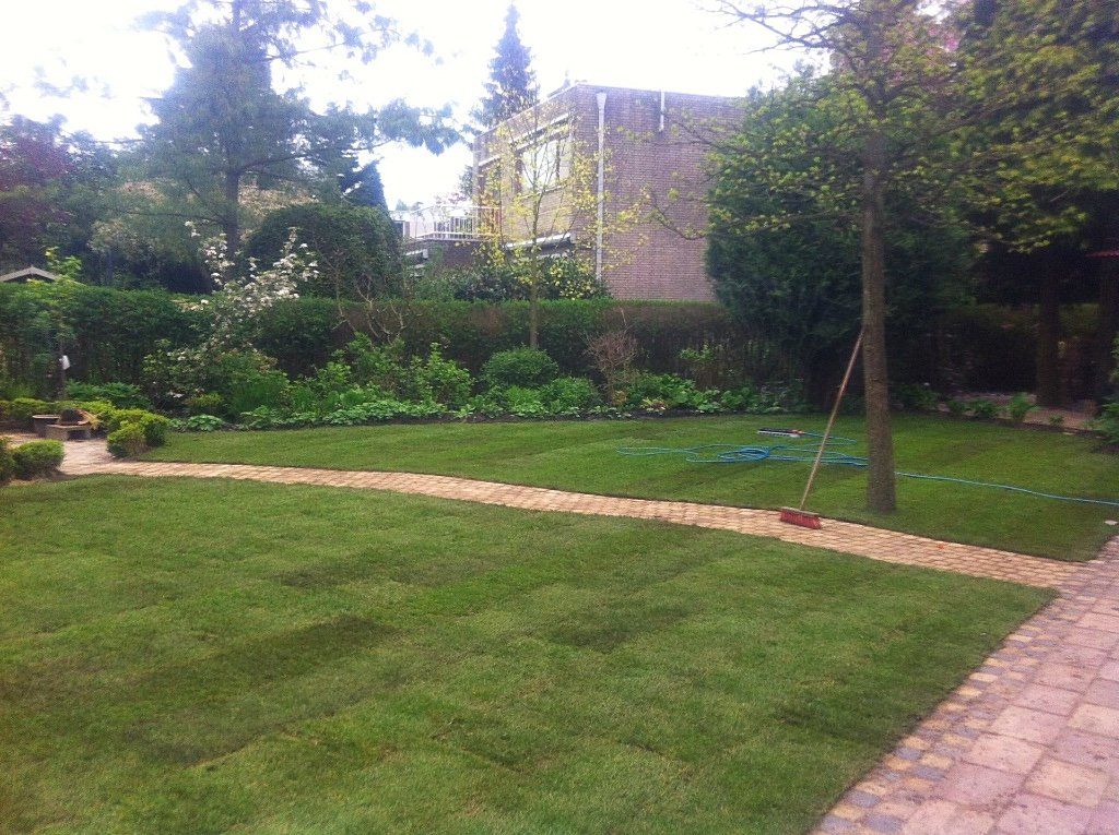 Een weelderig groen gazon met een gebogen bakstenen pad dat door een tuin leidt, met bomen en struiken op de achtergrond.