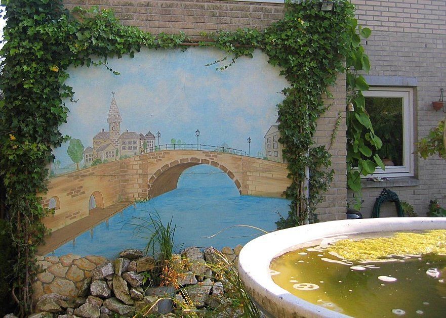 Muurschildering van een brug en gebouwen boven het water, omlijst door klimop op een stenen muur, met een vogelbad op de voorgrond.