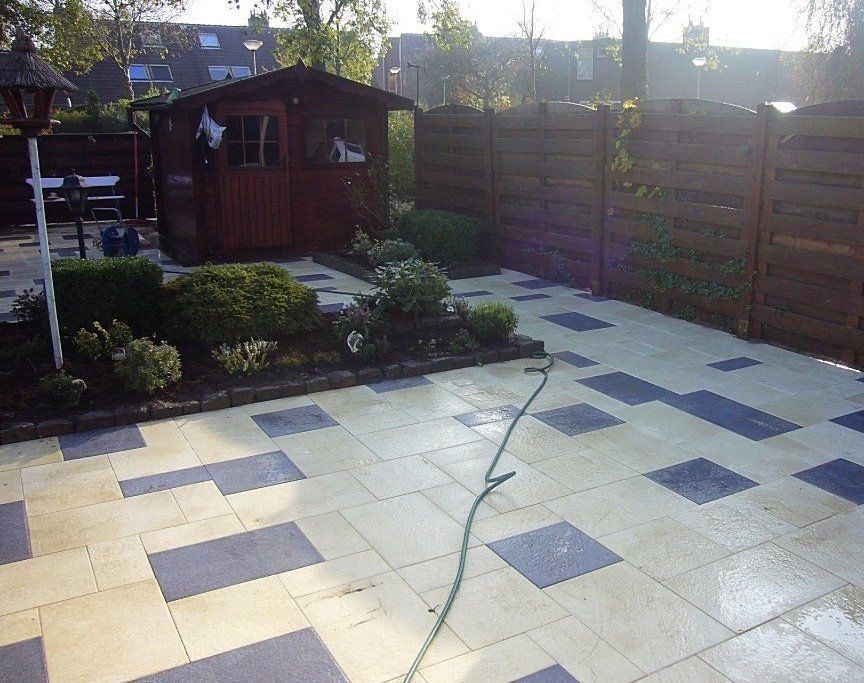 Terras met afwisselend beige en blauwe tegels, tuinschuur, houten schutting en groen.