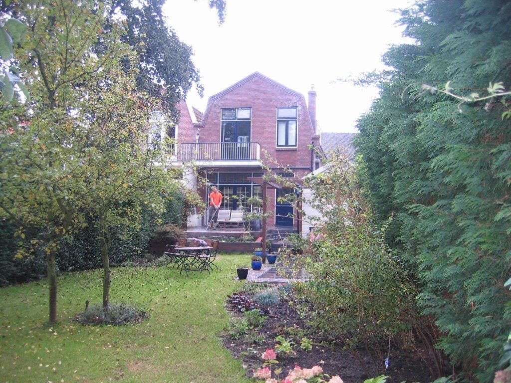 Achtertuin met gras, bomen, een bakstenen huis met een balkon en een terras met een tafel.