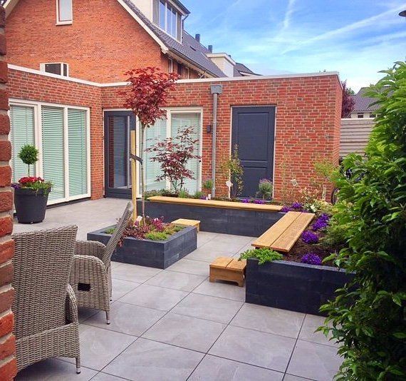 Een modern bakstenen terras met plantenbakken, zitgelegenheid en weelderig groen, naast een huis met grote ramen.