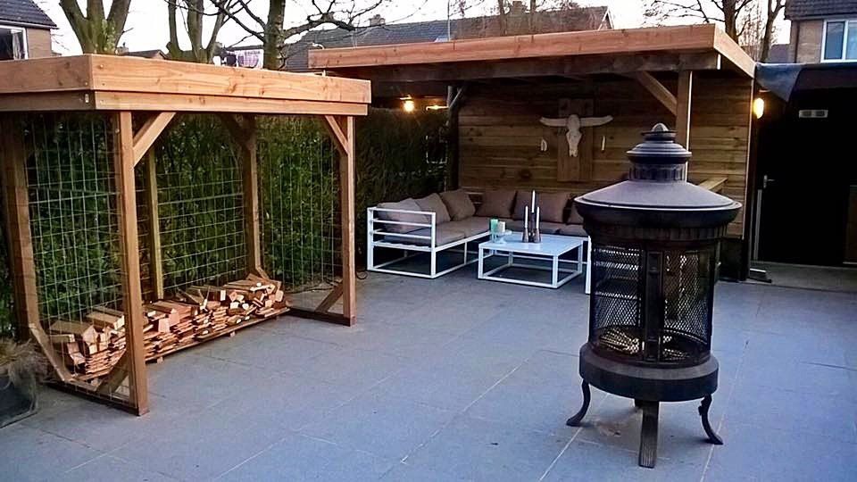 Buitenpatio met zitgedeelte onder een houten dak. Een vuurkuil, houtopslag en groen zijn zichtbaar.