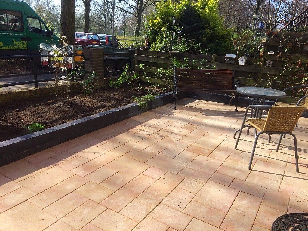 Terras met baksteenachtige bestrating, een klein bloembed en een tafel met stoelen. Een groene bestelwagen is gedeeltelijk zichtbaar.