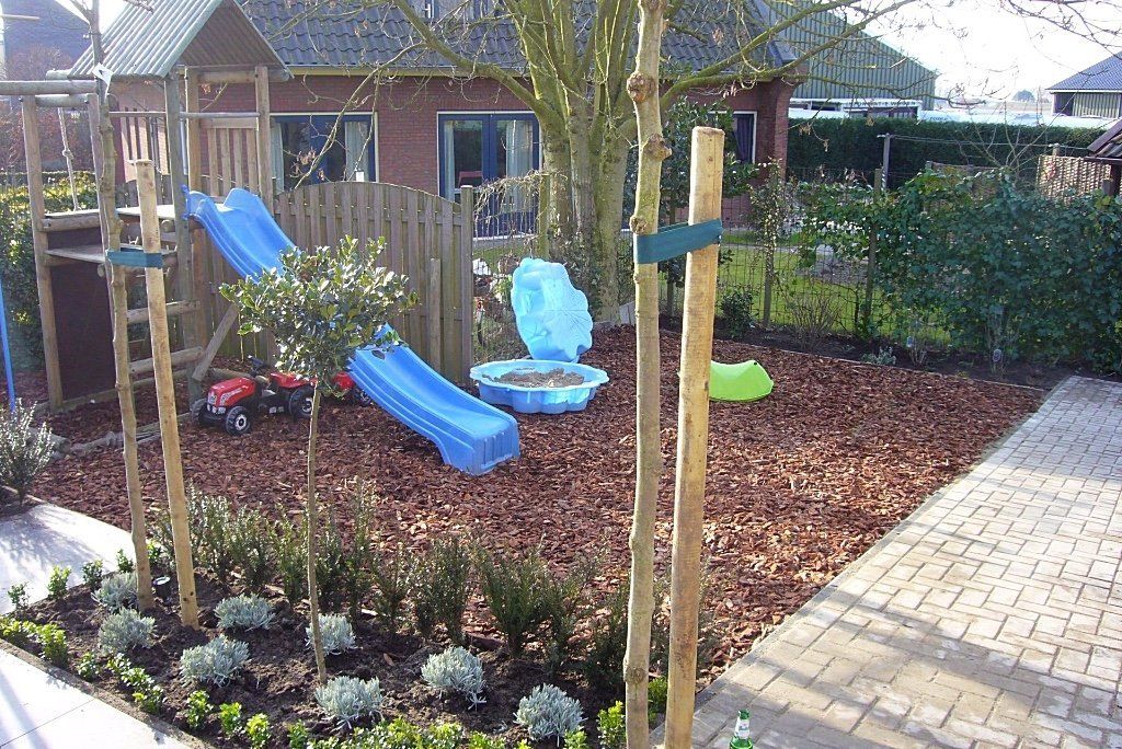 Speeltuin met glijbanen, houtsnippers en kleine plantjes in een achtertuin. Op de achtergrond is een gebouw te zien.