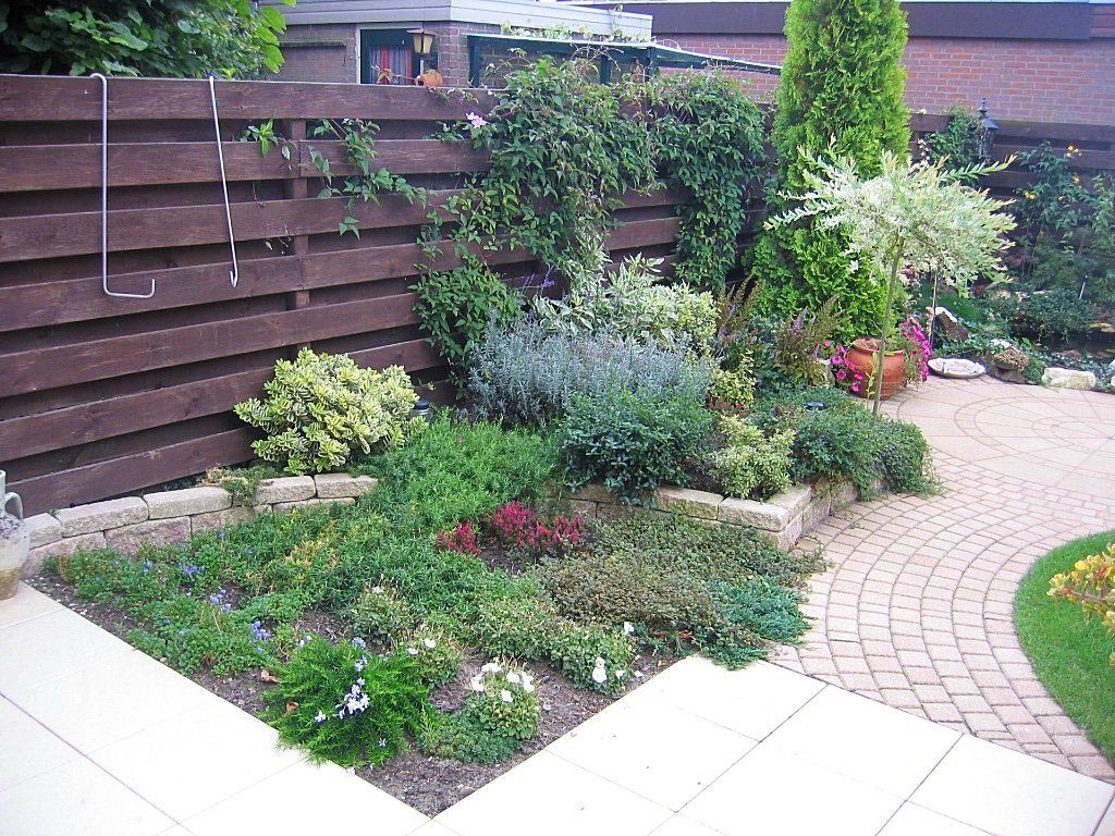 Een kleurrijk bloembed met diverse planten en een bruine schutting op de achtergrond; een beige bakstenen pad.