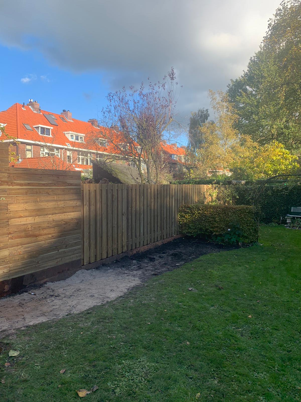 Een houten schutting begrenst een achtertuin met een keermuur en gebouwen op de achtergrond. Groen gras en bomen zijn zichtbaar.