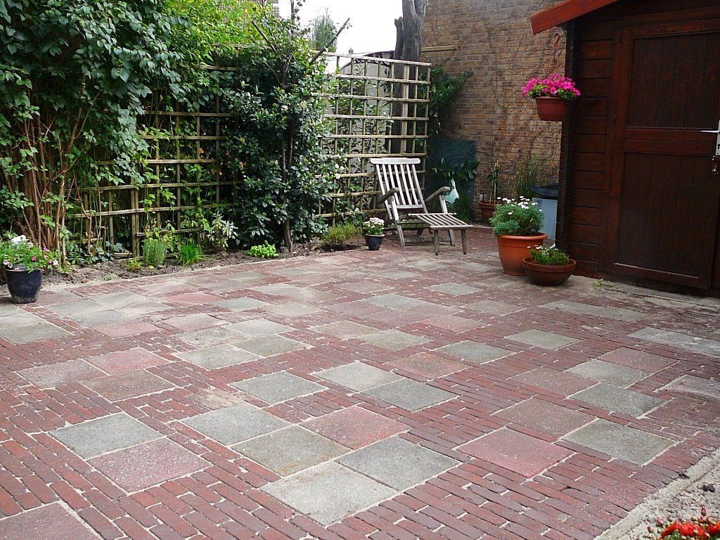 Een stenen terras met een tuin, een pergola, een stoel en een schuurtje met bloemen.